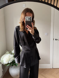 Woman wearing a black outfit taking a mirror selfie in a room with a vase of flowers. Satin black blouse with a pintuck waist from Styled Clothing paired with satin wide leg trousers.