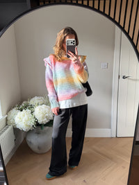 Woman wearing a colourful knit in a rainbow style gradient, laid over a boxy oversized tee and black satin trousers, taking a mirror selfie in a room with white flowers and a door.