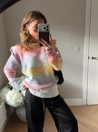 Woman wearing a colourful knitted jumper with ruffle shoulders taking a mirror selfie in a room with white walls and wooden flooring. Worn over a white tee and paired with black satin trousers.