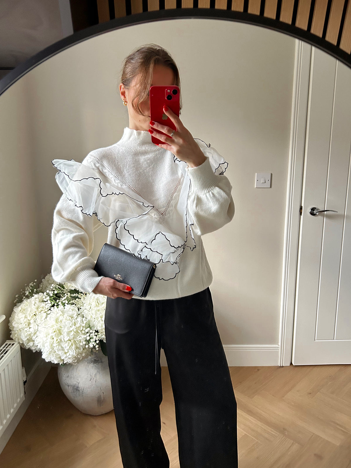 Person taking a mirror selfie wearing a white jumper with ruffled details and black satin trousers, holding a clutch bag.