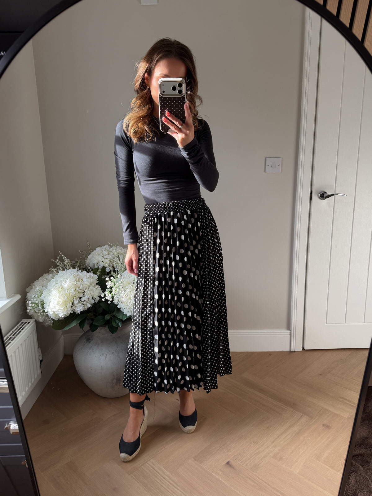 Mirror selfie of girl wearing a Black and White Polka Dot Skirt, midi length and elasticated waist. The main front panel of the pleated skirt is large white spots and the side panels are smaller polka dots.