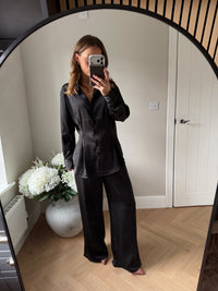 Girl wearing a black satin shirt with buttons paired with black trousers and heels taking a mirror selfie in a room with white walls and wooden floors.
