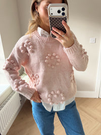 Woman wearing a pink knitted jumper with pom pom detailing in heart shapes. Paired over a white shirt and blue jeans.