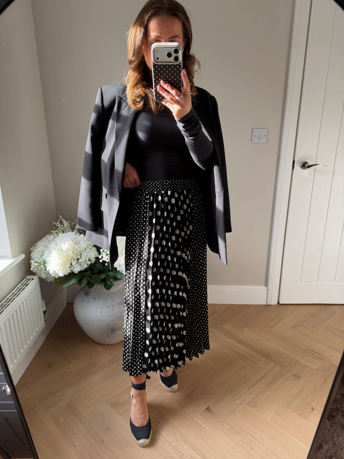 Woman wearing a black long sleeved top, oversized blazer over the shoulders and a white and black pleated polka dot skirt 