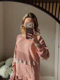 Person wearing a pink round neck jumper with pearl and tassel details.