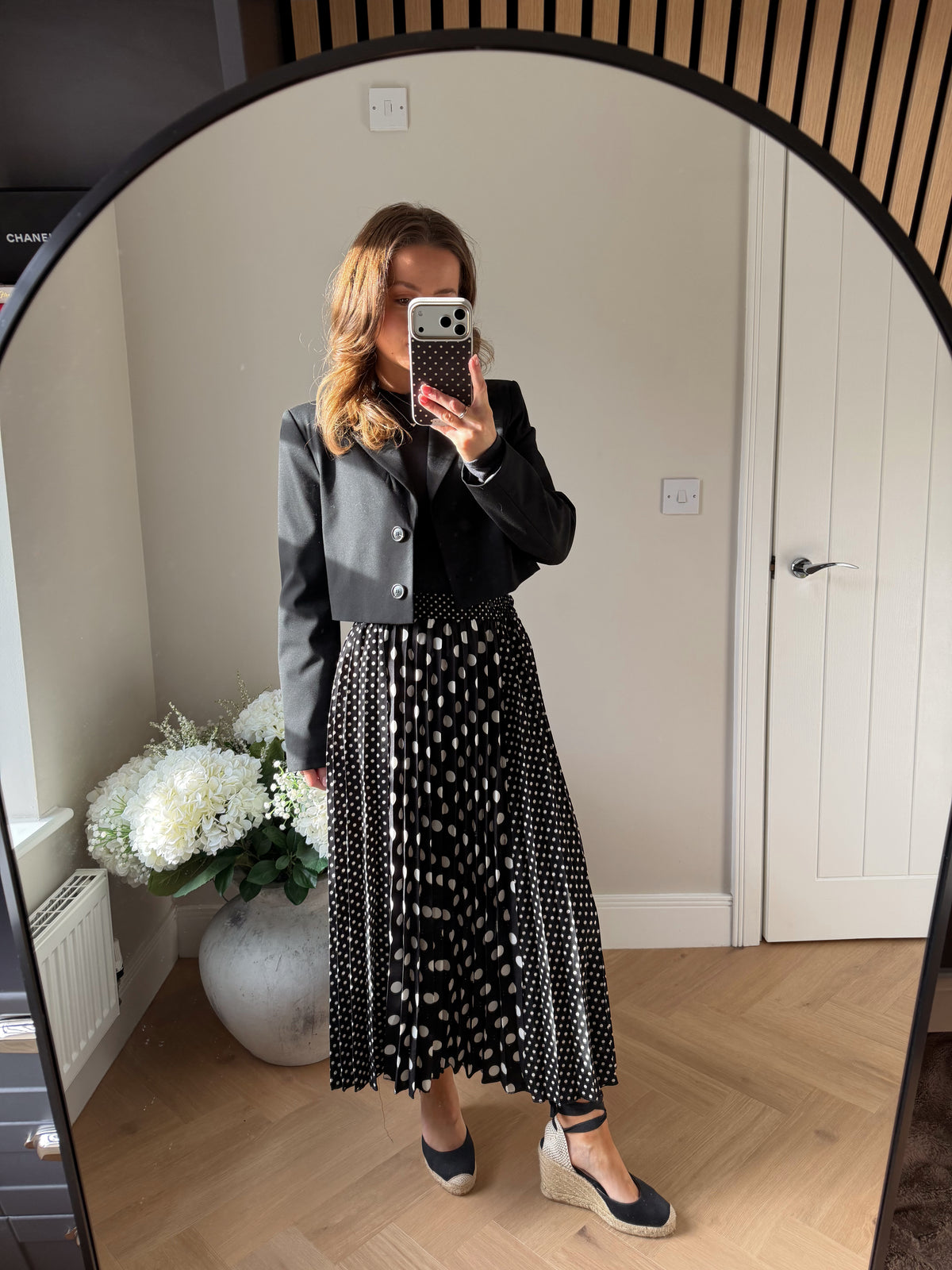 Woman taking a mirror selfie wearing a cropped black blazer jacket and polka dot skirt in a room with a white door and plant.