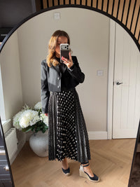 Woman taking a mirror selfie wearing a cropped black blazer jacket and polka dot skirt in a room with a white door and plant.