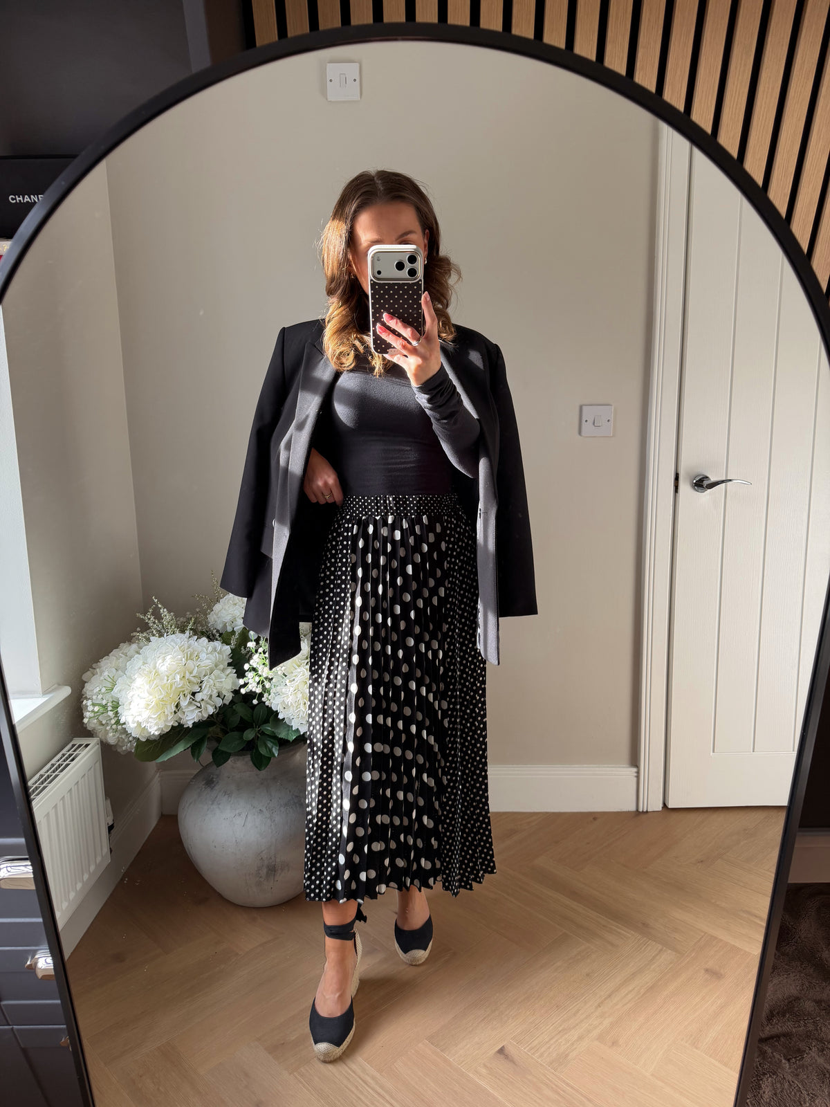 Woman wearing a black elasticated waist pleated skirt with front and back panels with large polka dots and smaller polka dots on the side panels. Paired with tie up summer espadrille wedges, long sleeve black top and oversized black blazer.