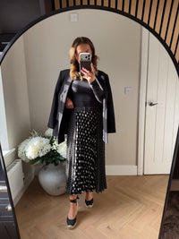 Woman wearing a black elasticated waist pleated skirt with front and back panels with large polka dots and smaller polka dots on the side panels. Paired with tie up summer espadrille wedges, long sleeve black top and oversized black blazer.