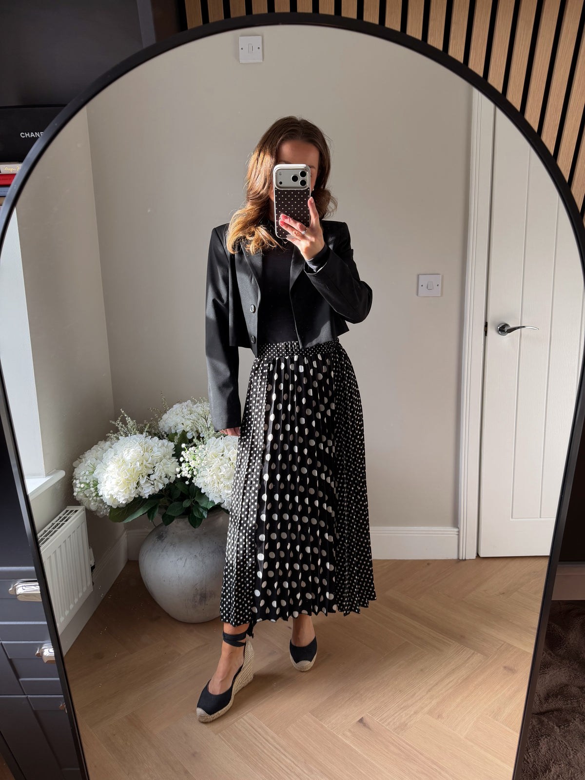 Mirror selfie of girl wearing a Black and White Polka Dot Skirt, midi length and elasticated waist. The main front panel of the pleated skirt is large white spots and the side panels are smaller polka dots. Paired with tie up black wedges and a cropped black blazer over a long sleeved black top.