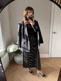 Person taking a mirror selfie wearing an oversized black blazer, black long sleeved  top, and patterned skirt. The skirt is midi length polka dot skirt with the background black with white spots. The skirt is pleated and the front and back sections are large polka dots and the side panels are smaller polka dots.